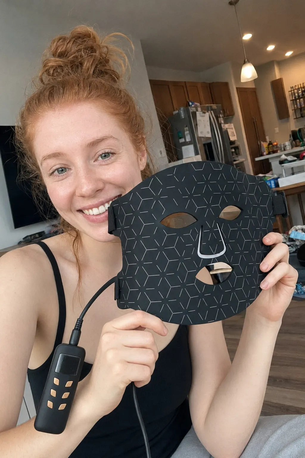 A smiling woman with red hair in a bun holds up a black LED facial mask with a geometric pattern and a connected controller, set against a blurred home interior background.