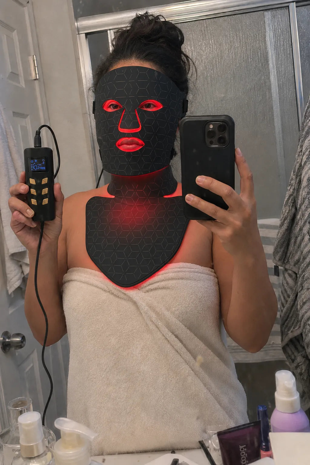 A woman takes a mirror selfie in a bathroom while wearing a black LED light therapy face and neck mask that glows red around the eyes, mouth, and neck. She is wrapped in a beige towel and holds a black handheld controller in one hand and a black smartphone in the other.