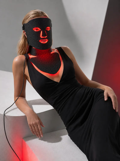 A woman reclining on white geometric steps, wearing a black LED light therapy face mask that glows red around her eyes, nose, and mouth, along with a matching black dress.