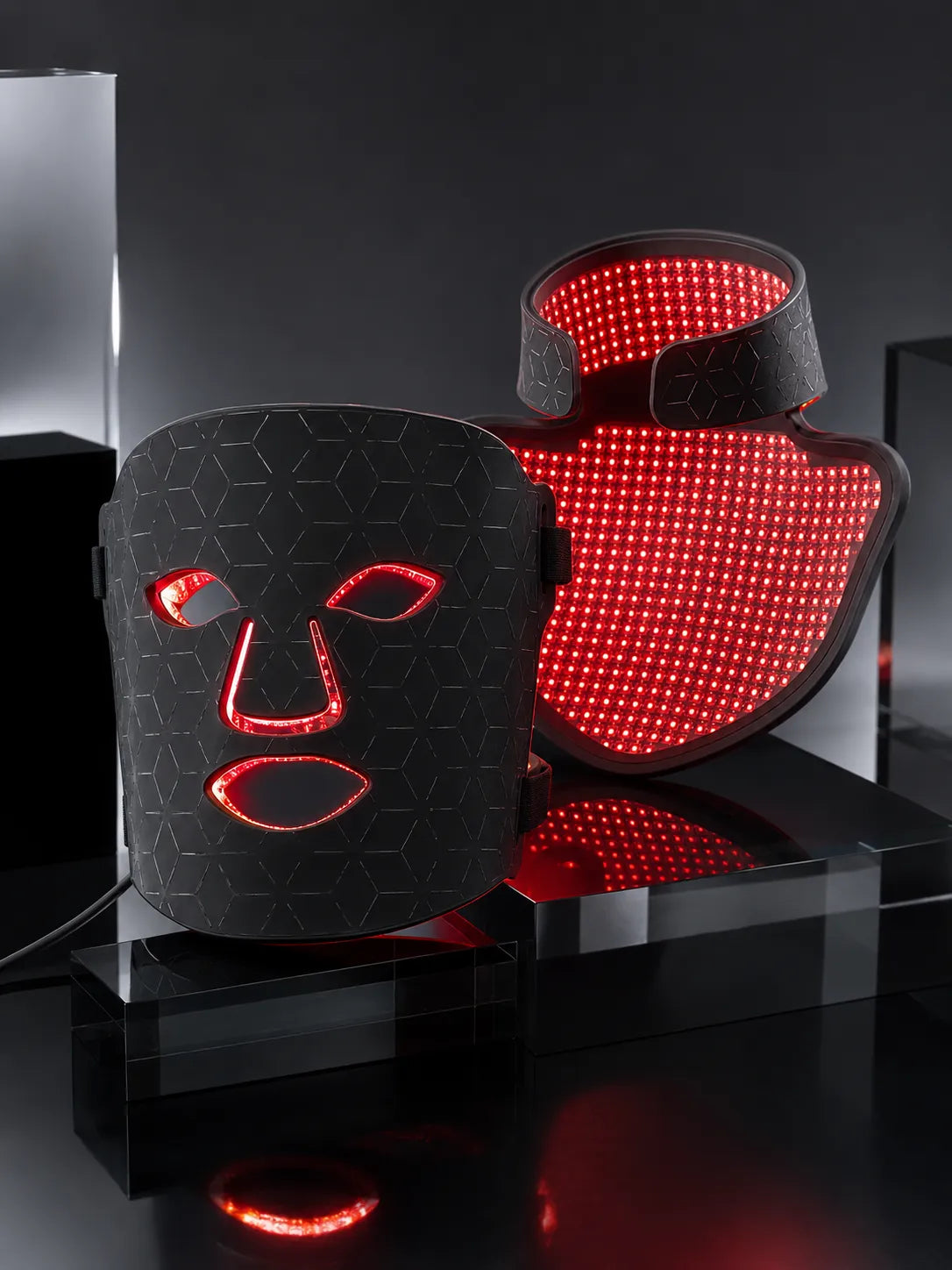 Two black LED light therapy masks displayed on a glossy black surface against a dark studio background. One mask is shown closed with a hexagonal pattern, and the other is open, revealing bright red illuminated light points inside.