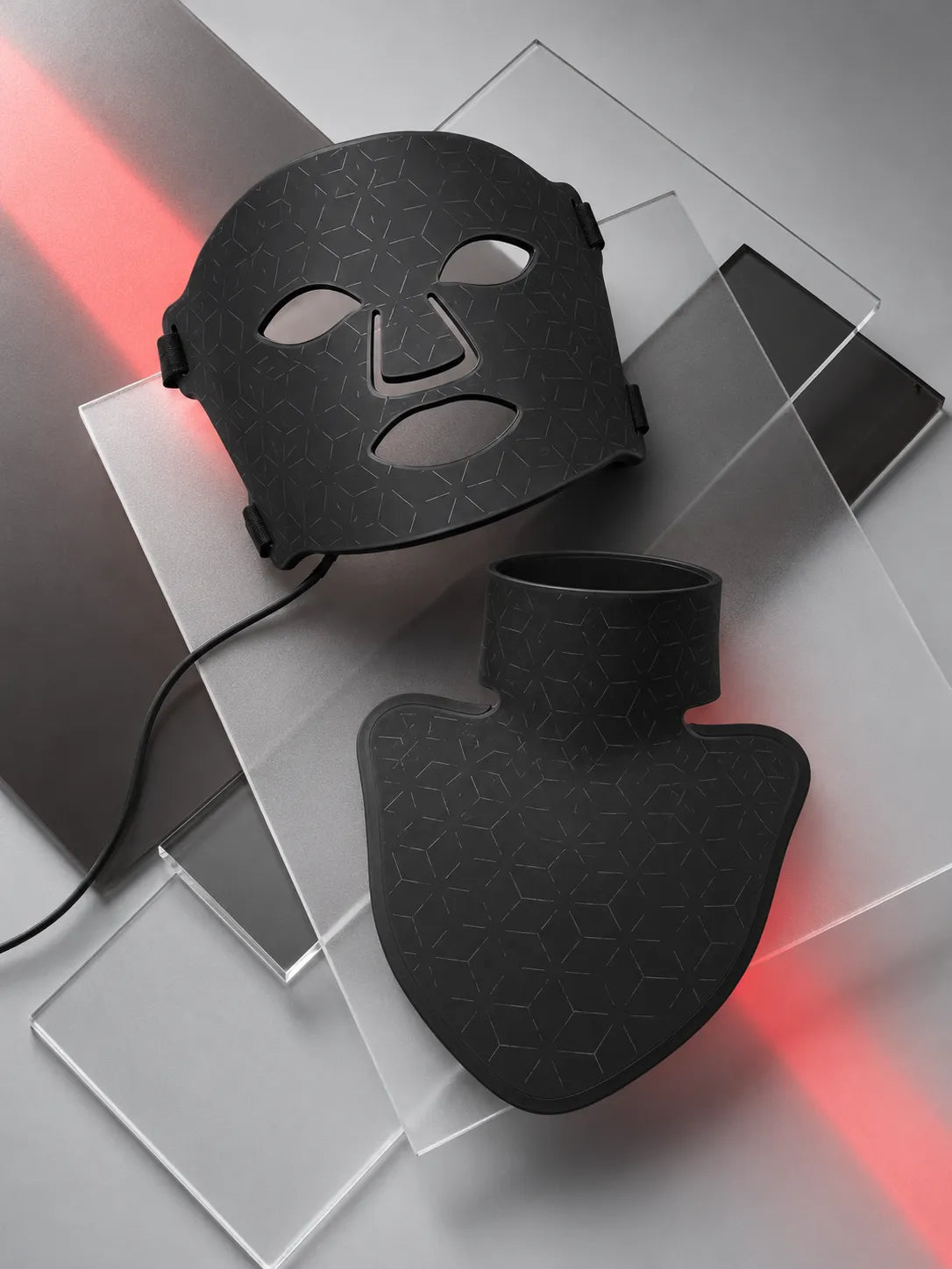 Black LED light therapy face and neck masks with a subtle geometric pattern, displayed on clear acrylic blocks with red light reflecting off the surface.