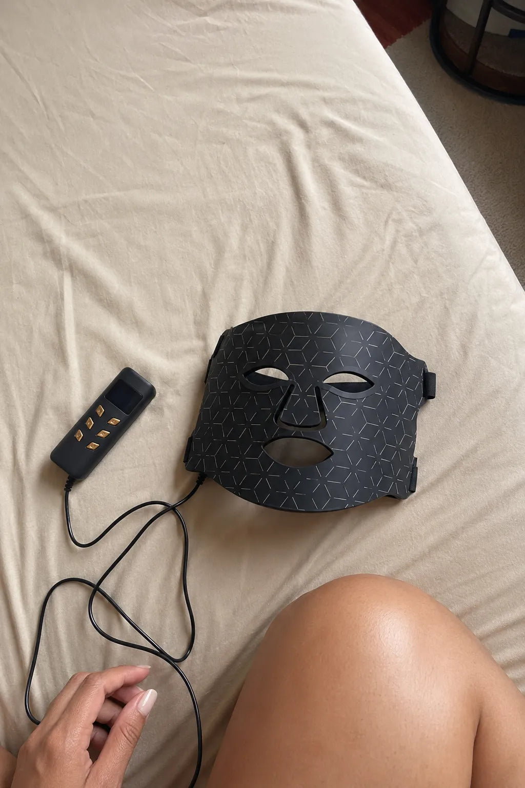A black LED facial therapy mask with a geometric pattern, shown next to its black remote control on a beige bed sheet. A person's hand and leg are visible in the foreground.